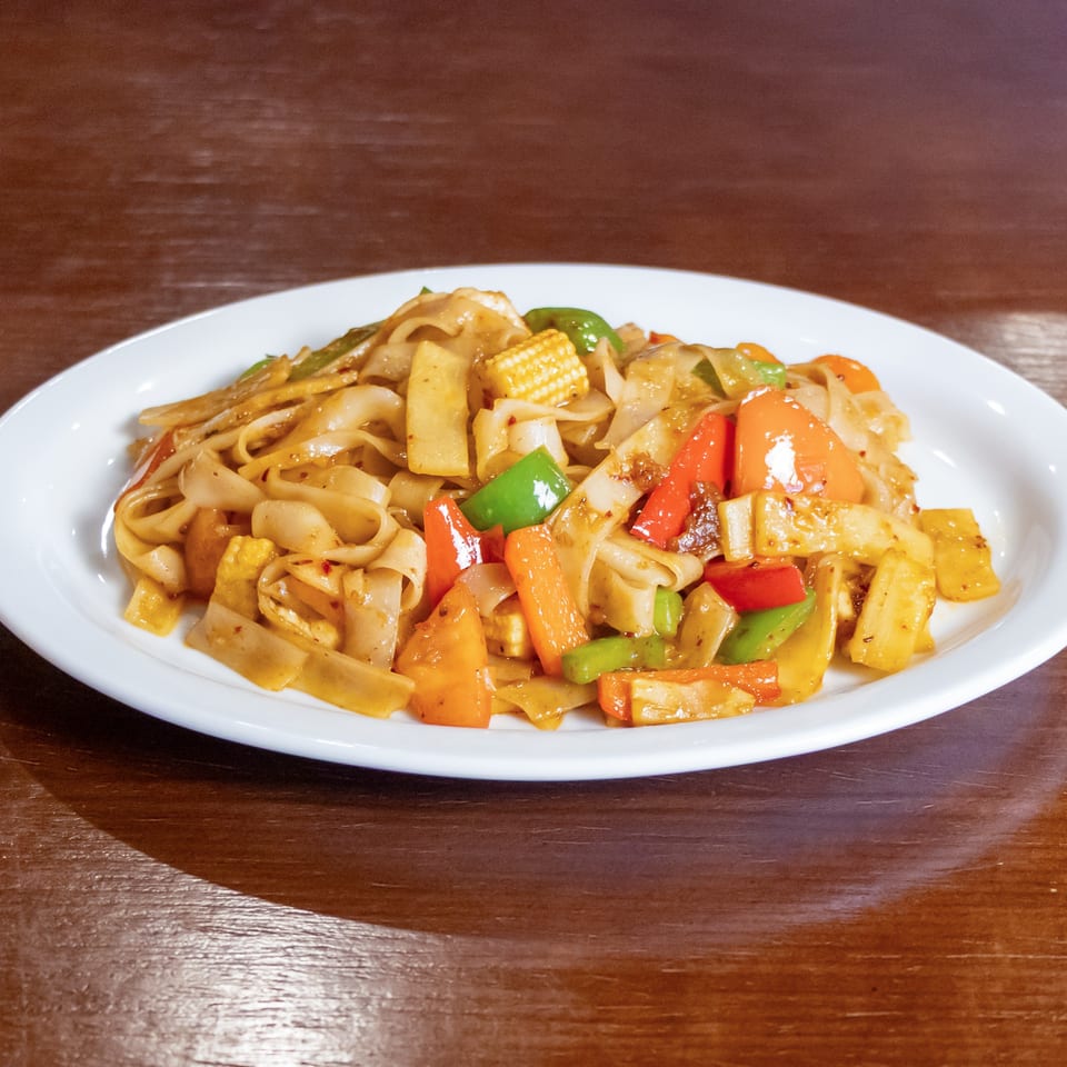 Plate of stir-fried flat noodles with colorful bell peppers, baby corn, carrots, and other vegetables on a wooden table.